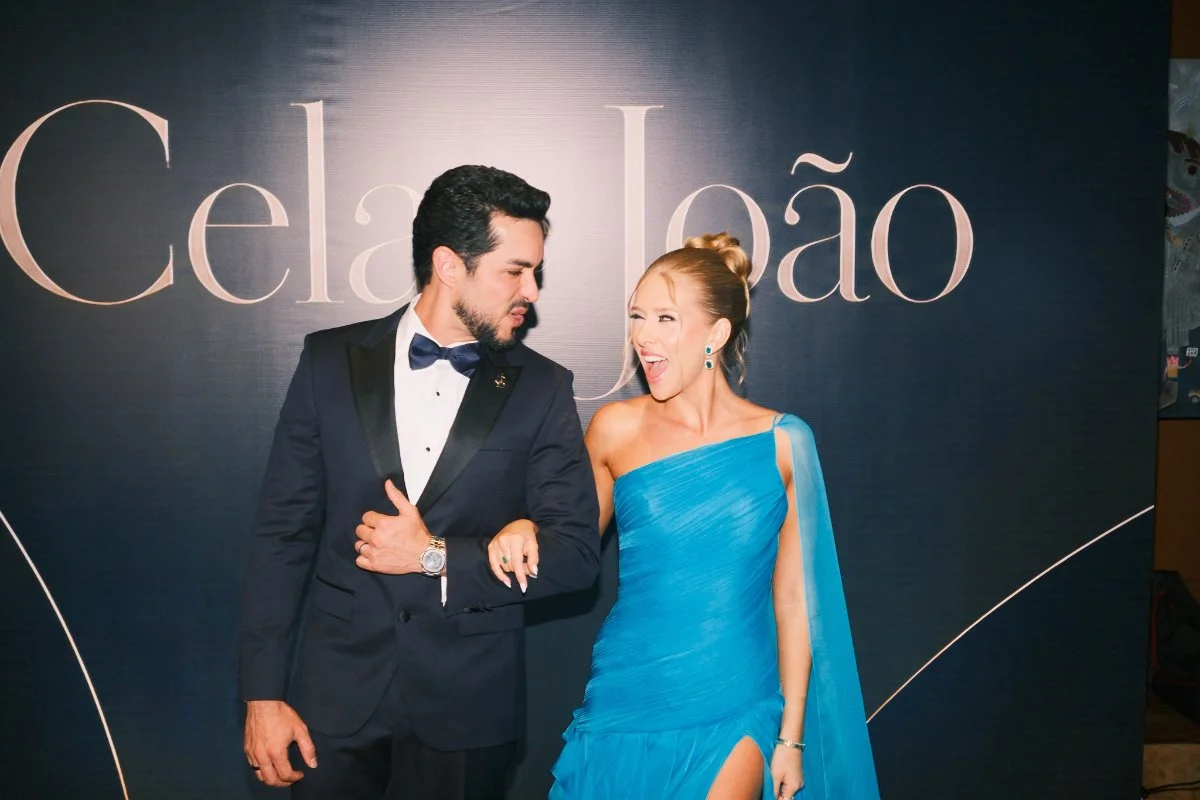 Cela Lopes e Jo&atilde;o Brasio de bra&ccedil;os dados em frente ao painel da festa de anivers&aacute;rio